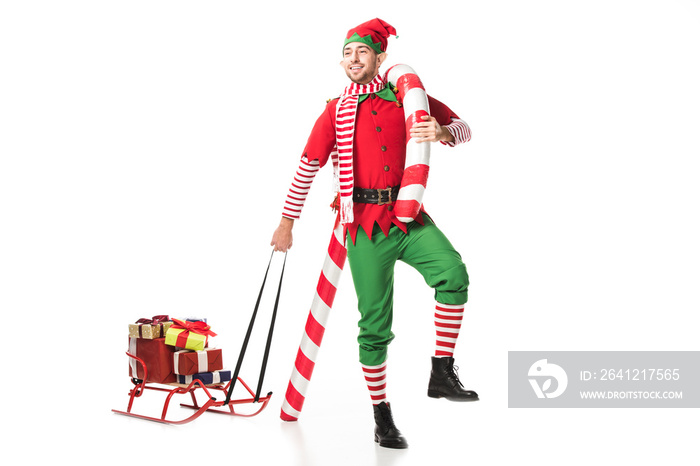 happy man in christmas elf costume carrying sleigh with presents and big candy cane isolated on whit