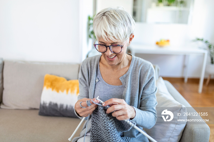 Elderly woman in glasses sit on couch at home smile holding knitting needles and yarn knits clothes 