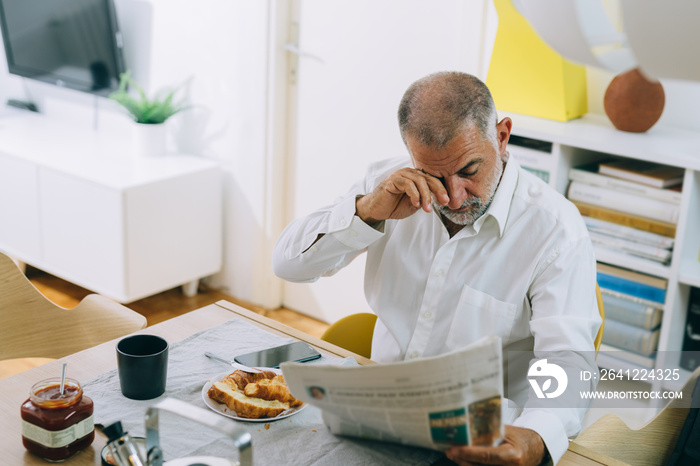 middle aged man reading newspapers while having breakfast at home, having eyes problem