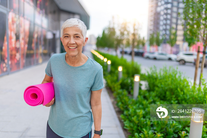 An elderly woman poses for a portrait after her workout. Senior woman walking with a yoga mat outsid