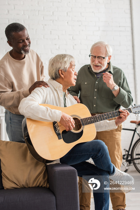 Cheerful interracial men standing near asian friend playing acoustic guitar at home.