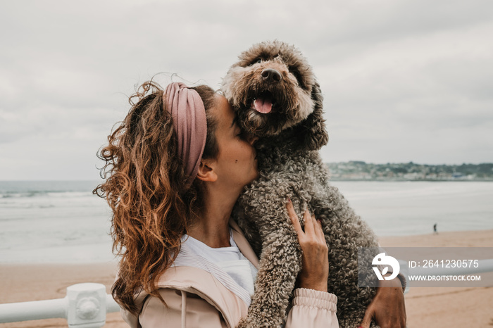 。一名身穿粉色外套的年轻女子，在海滨与她可爱的棕色西班牙水犬玩耍