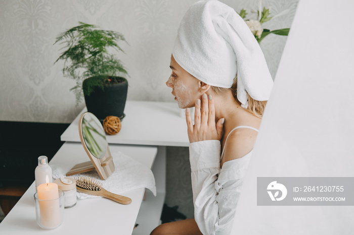 Young woman in white towel chilling in bedroom and making clay facial mask near mirror. Girl doing b