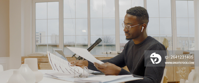 Handsome African American male working from home, using laptop in living room, checking documents