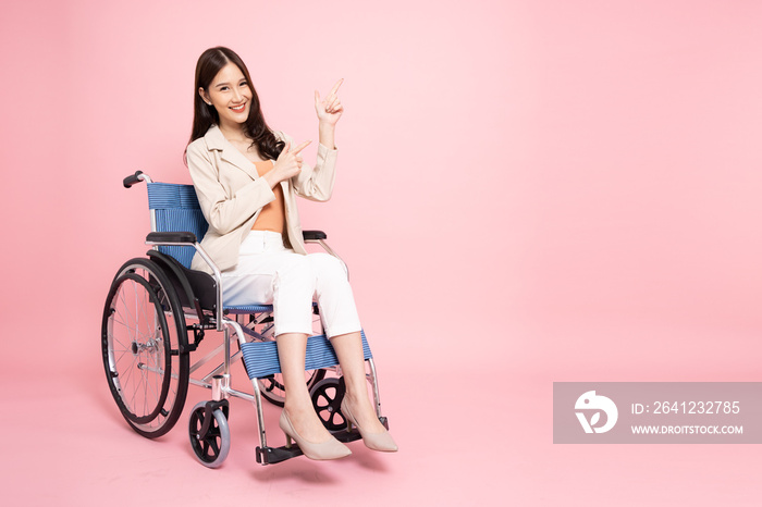 Smiling young Asian businesswoman sitting on wheelchair and pointing to empty copy space isolated on