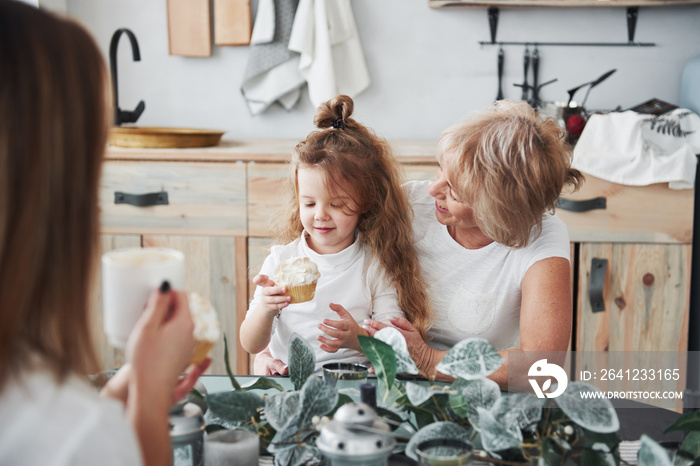 Drinking coffee and watching at kid. Mother, grandmother and daughter having good time in the kitche