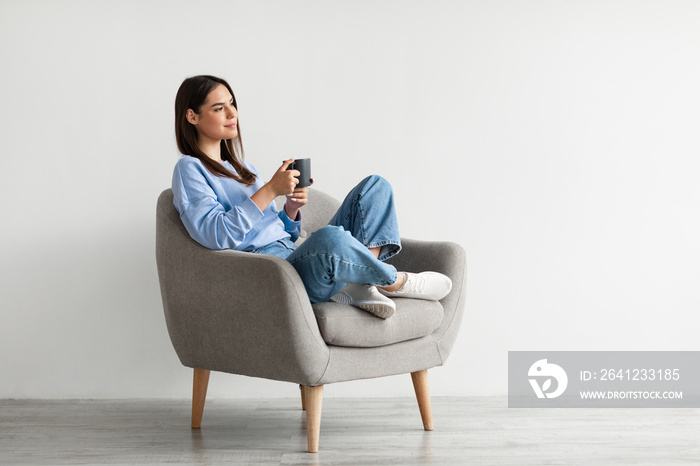 Positive young lady sitting in armchair with crossed legs, warming hands on cup of hot coffee, enjoy