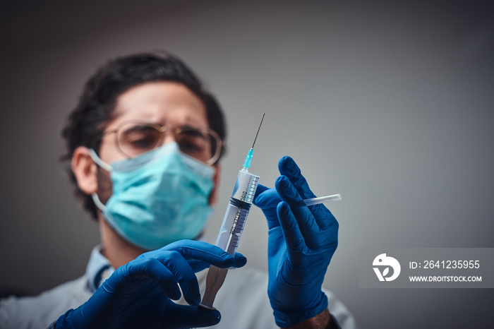 Focused doctor in protective mask and gloves is preparing a remedy in syringe.