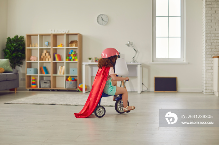 戴着头盔和红色斗篷的学龄前女孩在舒适的托儿所里骑着三轮车