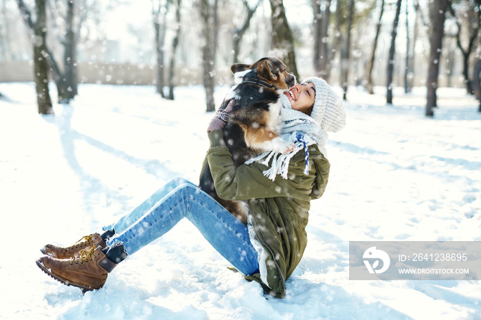 戴着羊毛帽、戴着温暖的长围巾的年轻快乐女人在雪地里的冬季公园里抱着她的宠物。