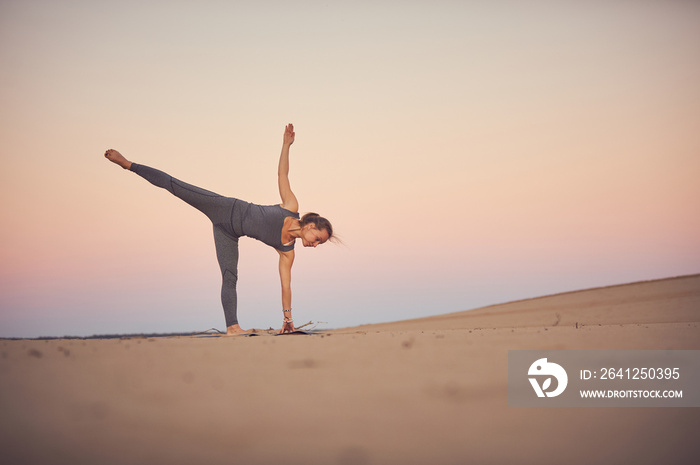 Beautiful young woman practices yoga asana Ardha Chandrasana - Half Moon pose in the desert at sunse