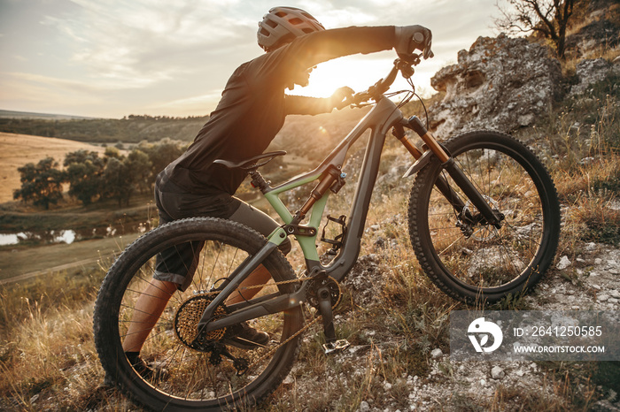 Young male cyclist rolling bike on grassy mountain slope in sundown