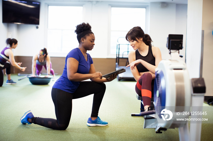 Instructor showing tablet computer to female athlete exercising on rowing machine at gym