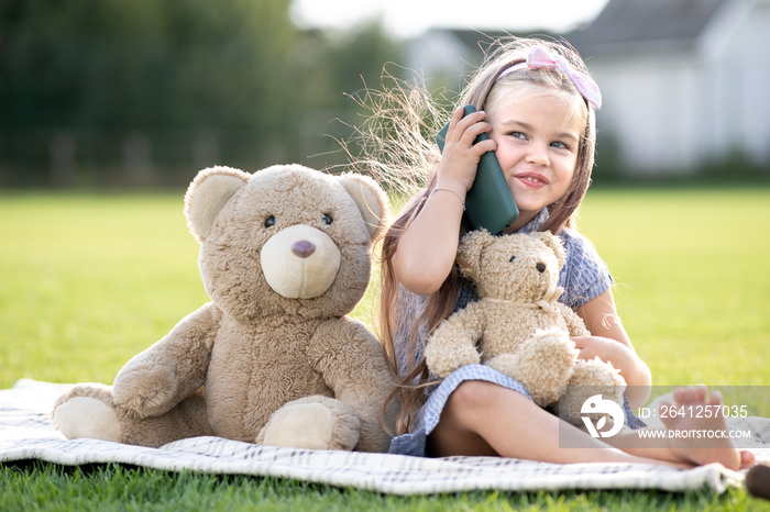 漂亮的小女孩坐在绿草草坪上的避暑公园里，拿着她的泰迪熊玩具在mobi上说话
