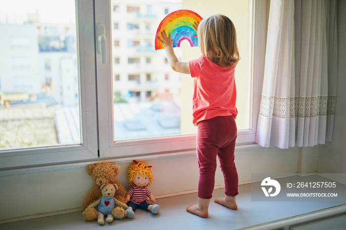 Adorable toddler girl attaching drawing of rainbow to window glass