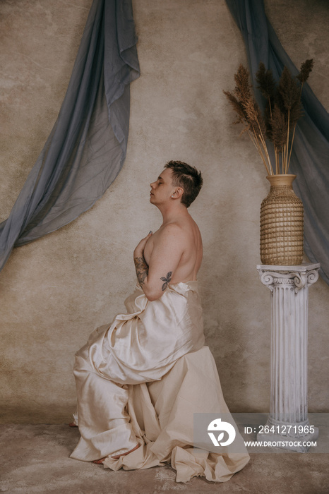 profile of a shirtless non-binary person sitting on pedestal with hands on chest eyes closed