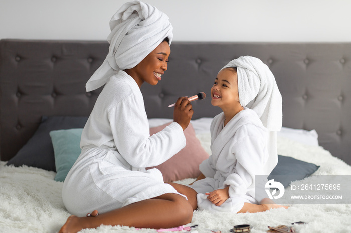 Happy black mom and her cute daughter wearing bath robes and towels, applying makeup on family spa d