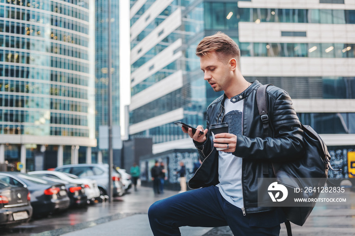 Attractive stylish young man in a leather jacket with a smartphone and take-out coffee in his hands 