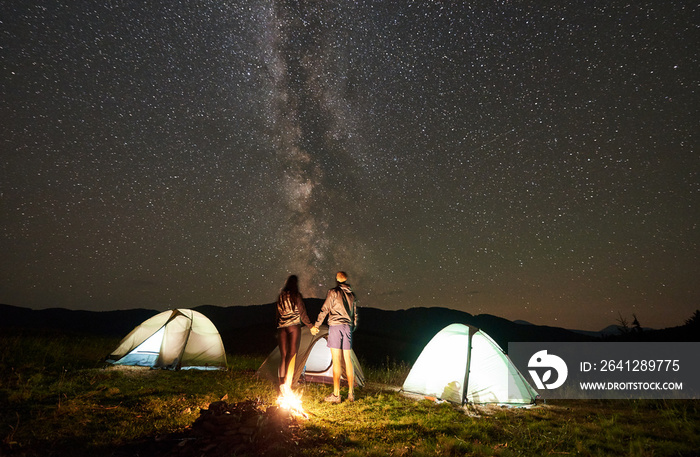 Active couple hikers resting at summer night camping in the mountains. Back view of man and woman st