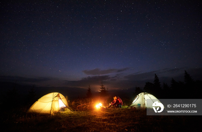 Camping in mountains at night. Bright bonfire burning between two tourists, boy and girl sitting opp