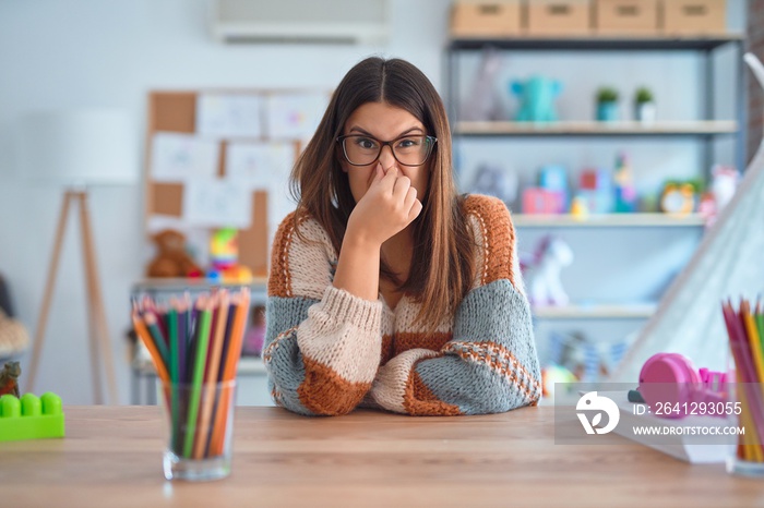 Young beautiful teacher woman wearing sweater and glasses sitting on desk at kindergarten smelling s