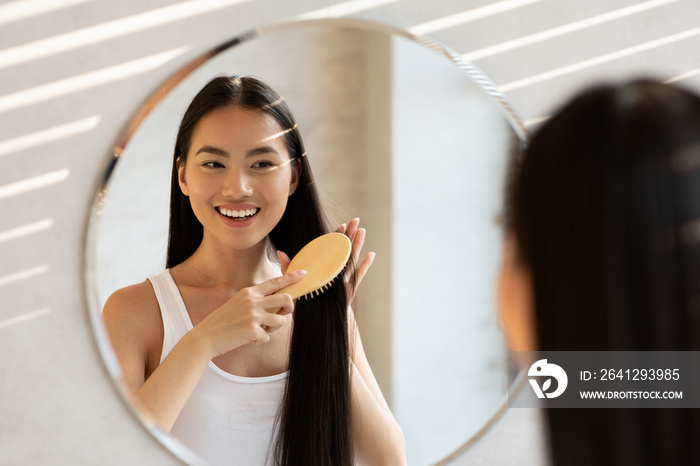 Cheerful asian lady brushing hair, standing in front of mirror