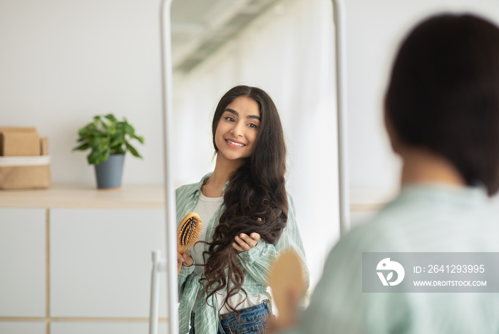 Cheerful Indian woman brushing her long beautiful hair with wooden brush in front of mirror at home