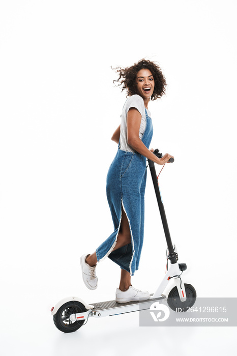 Image of excited african american woman dressed in denim overalls smiling at camera while riding on 