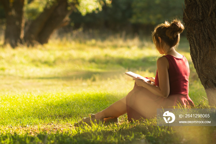 sunny portrait of a beautiful girl sitting on green glade under an arch of tree branches with book, 