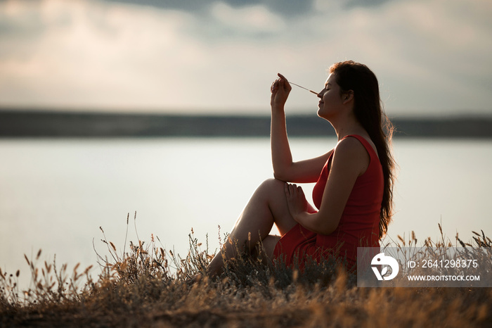 Smiling beautiful girl with blade of grass that touching her nose, sitting near the water on the gra