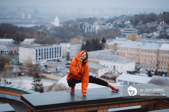 Girl in an orange jacket poses on the roof of a building in the city center