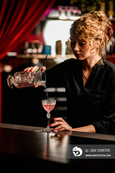 view of beautiful female bartender pouring cocktail from mixing cup into crystal glass