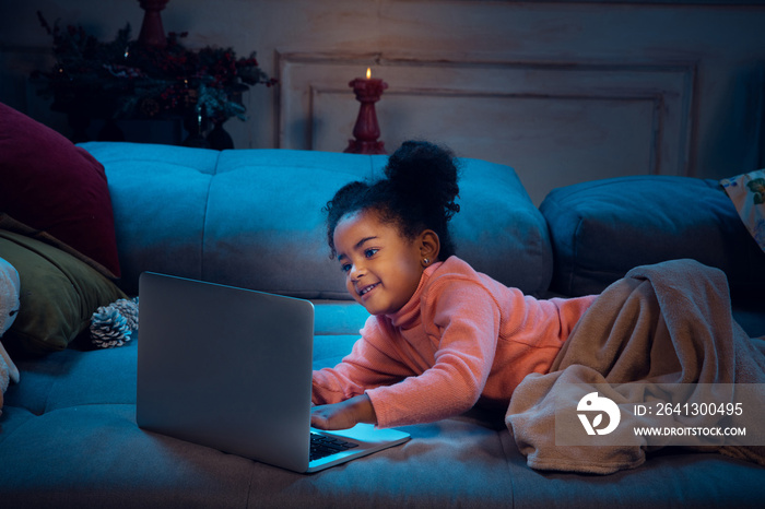 Happy african-american little girl during video call with laptop and home devices, looks delighted a