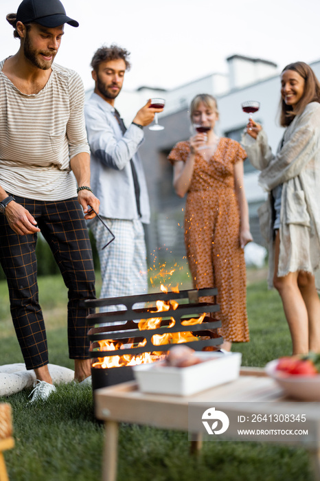 Friends have a picnic, making fire at barbeque, talking and drinking wine on the green lawn at backy