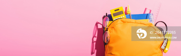 horizontal image of yellow backpack packed with school supplies and eyeglasses on pink