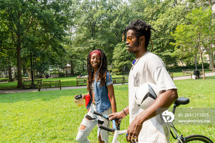 Stylish young people walking on lawn in park with bicycle