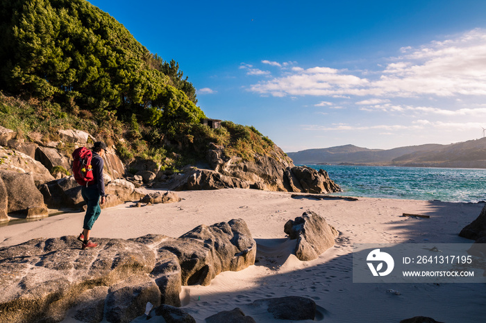 Hiker girl with backpack rests standing and looks at the turquoise sea on a small rocky beach of the