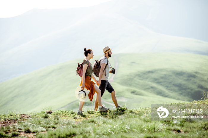 Beautiful couple walking with backpacks on the green meadow, while traveling in the mountains during