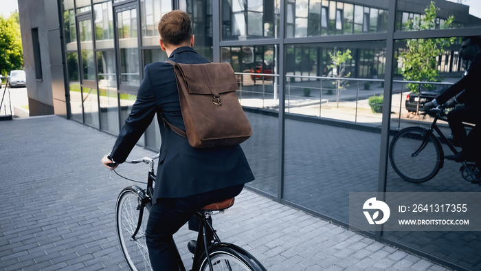 back view of businessman in suit with leather brown backpack riding bicycle near building.