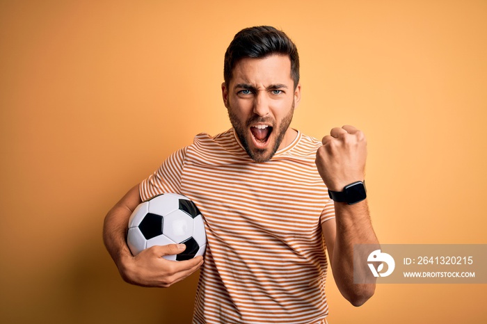 Handsome player man with beard playing soccer holding footballl ball over yellow background angry an