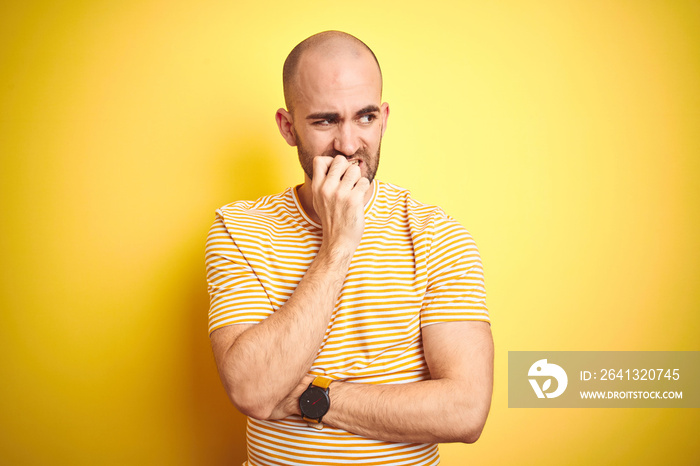 Young bald man with beard wearing casual striped t-shirt over yellow isolated background looking str