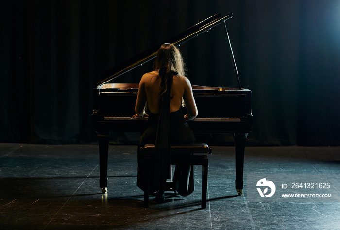 A woman from behind dressed in a black dress playing a black grand piano with the lid raised on a st