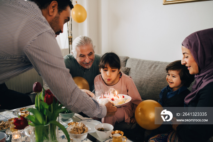 Happy family looking at girl blowing out candles on birthday cake at home