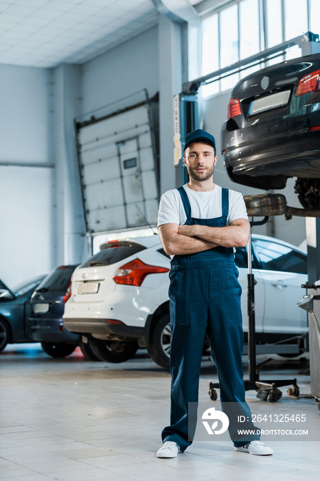 handsome car mechanic standing with crossed arms near cars