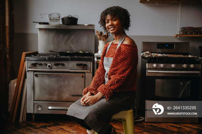 Portrait of smiling female student with curly hair sitting on stool against appliances in cooking cl