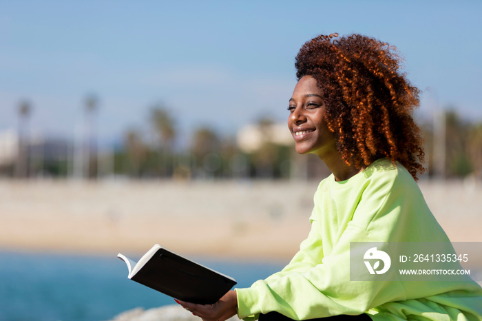 Side view of young curly afro woman sitting on a breakwater holding a book while smiling and looking