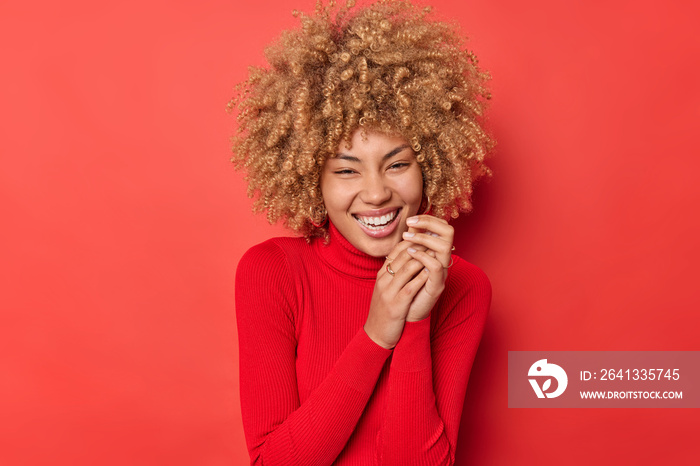 Horizontal shot of happy carfree woman with curly hair giggles positiely keeps hands together smiles