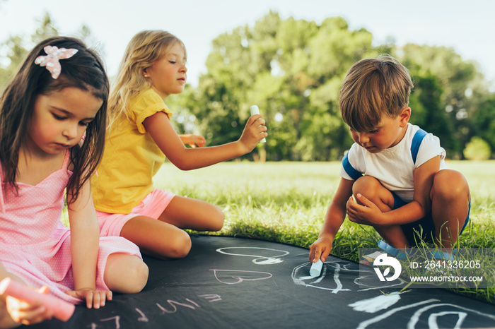 Outdoors image of kids sitting on the green grass playing with colorful chalks. Happy children drawi