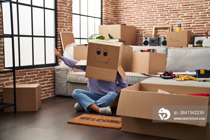 Young african american woman sitting on the floor with funny cardboard box on head at new home