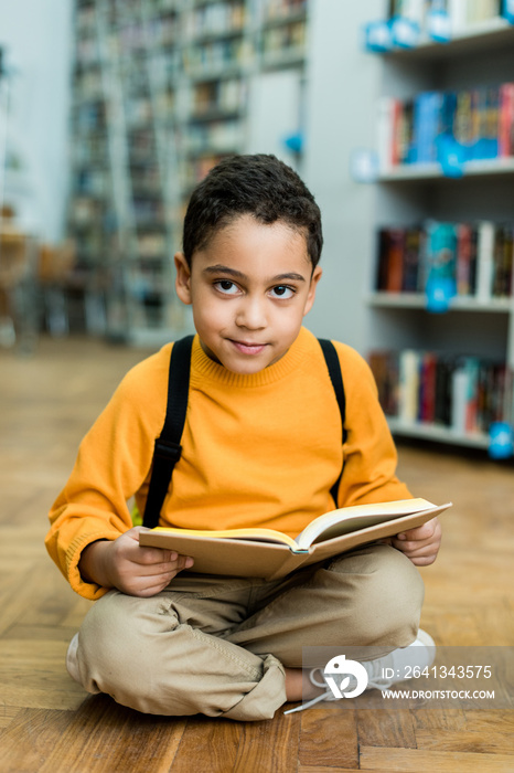 cheerful african american boy sitting on floor and holding book